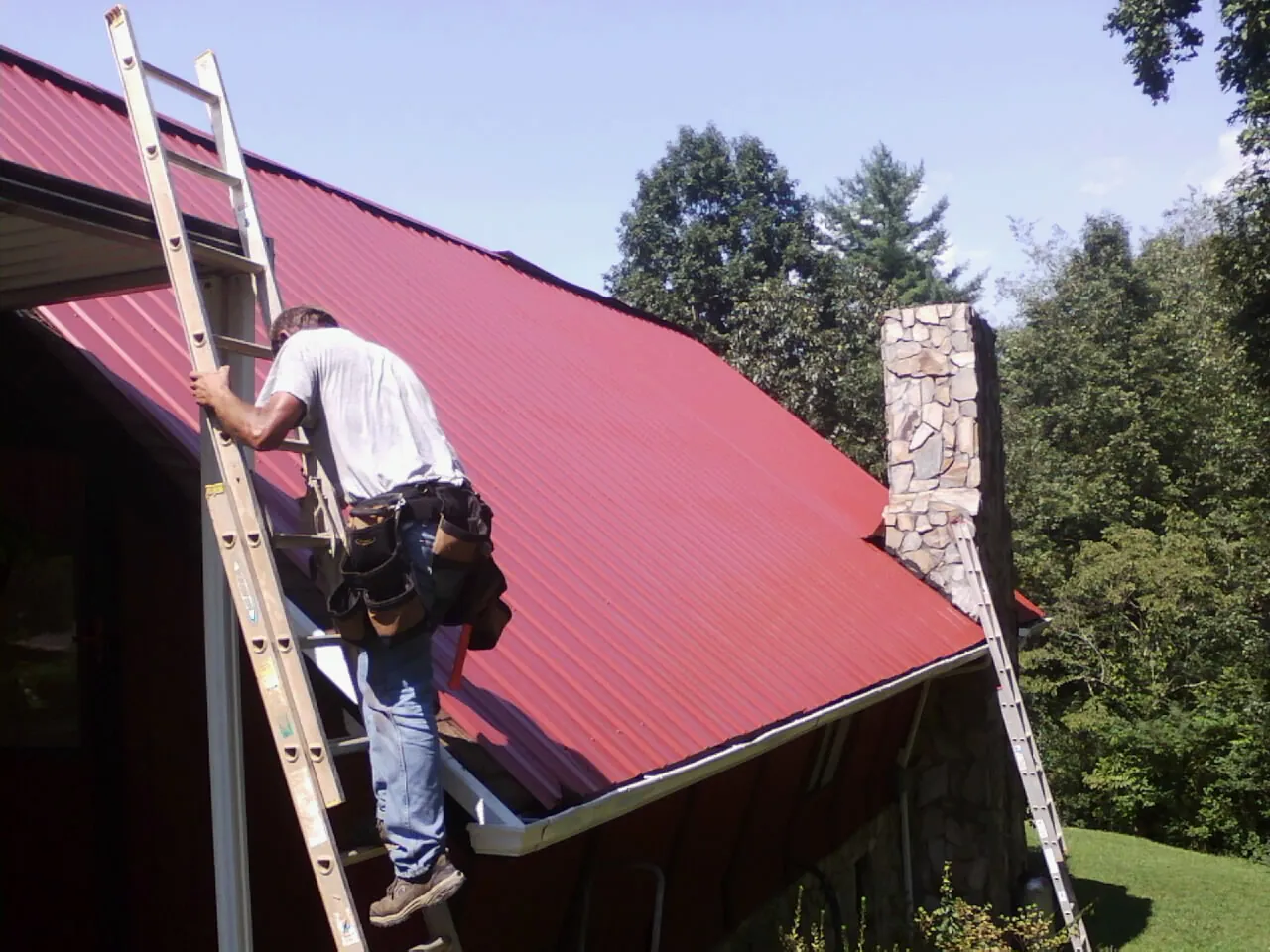 Roofer on a ladder beside a red metal roof and stone chimney — Soffit & Fascia Repair by Roofing Brothers in Nanticoke